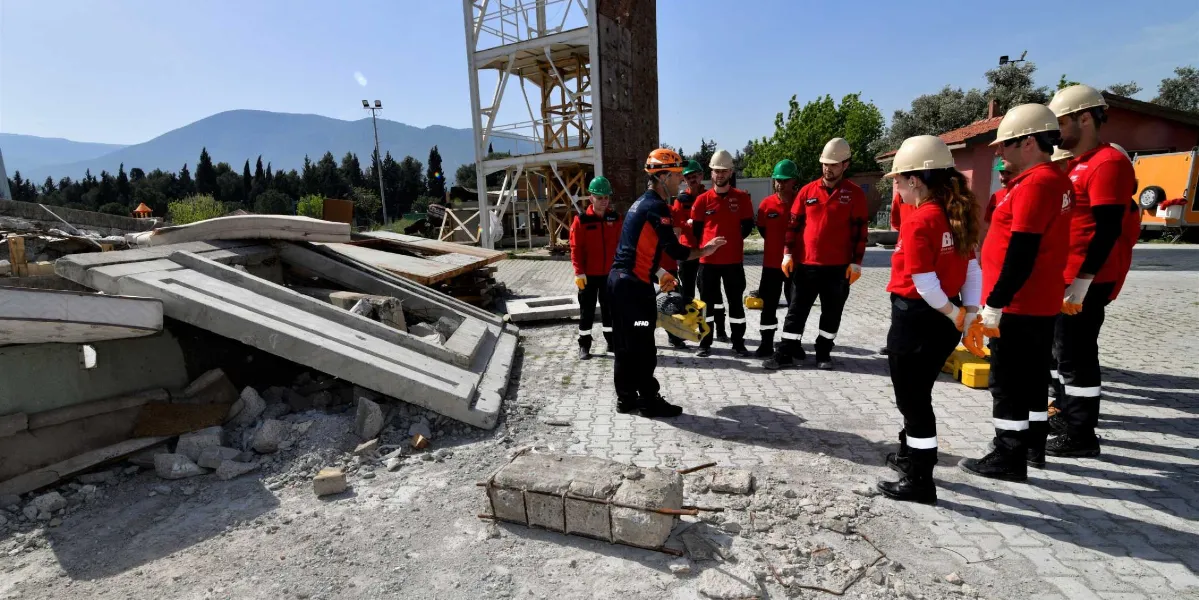 Açık havada, enkaz simülasyonu alanında eğitim alan Bornova Belediyesi arama kurtarma ekibi. Kırmızı tişörtlü ve baretli ekip üyeleri, ellerinde ekipman tutan bir eğitmeni dikkatle dinliyor. Arka planda metal bir eğitim kulesi ve dağ manzarası görülüyor.