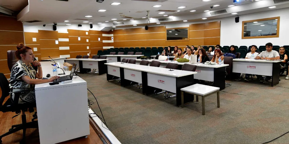 Modern bir konferans salonunda, kürsüde oturan bir konuşmacı ve onu dinleyen Peyzaj Mimarlığı öğrencileri yer alıyor. Salonun ön tarafında beyaz masalar ve arkada kademeli oturma düzeni bulunuyor. Masaların üzerinde Bornova Belediyesi logosu dikkat çekerken, öğrenciler sıralarda oturarak sunumu takip ediyor.