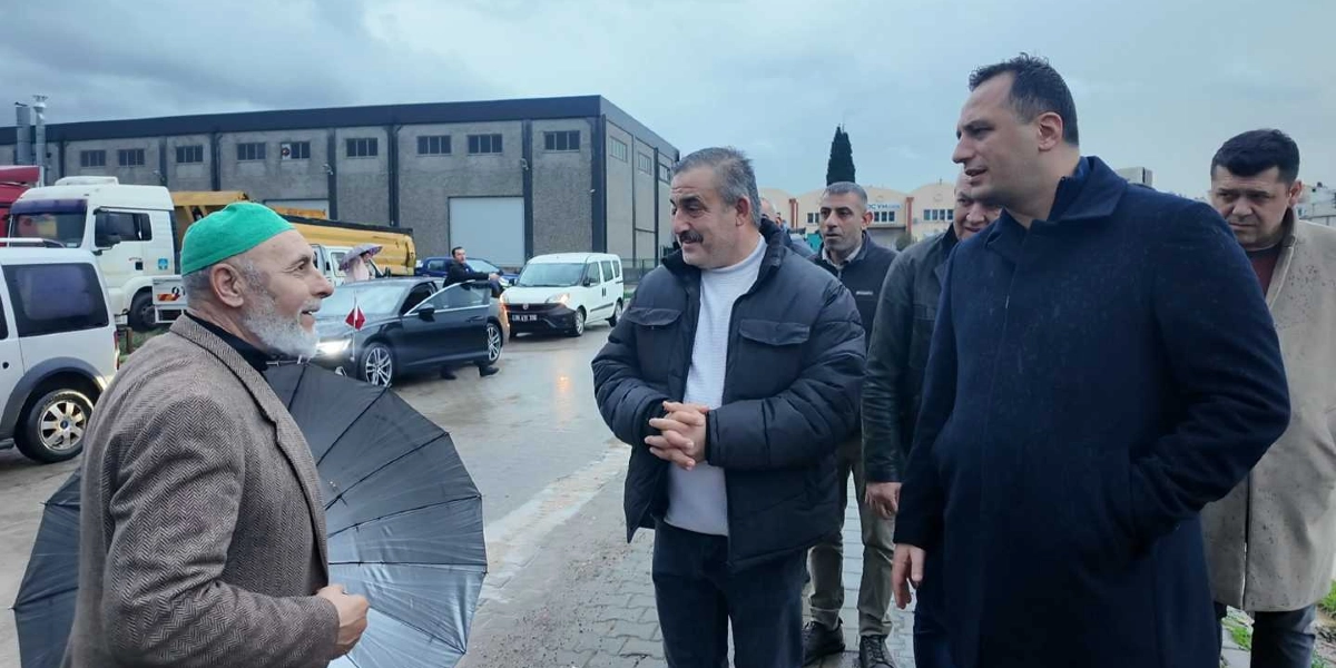 Bornova Belediyesi yetkilileri, Doğanlar mahallesindeki bir çalışma alanında yerel halkla bir araya gelerek incelemelerde bulunuyor. Açık havada, gri bir gökyüzü altında gerçekleşen buluşmada, yeşil takkeli ve sakallı bir vatandaş elinde şemsiyesiyle yetkililerle sohbet ediyor. Arka planda ise sanayi tipi binalar, park halindeki araçlar ve bir tır görülüyor.