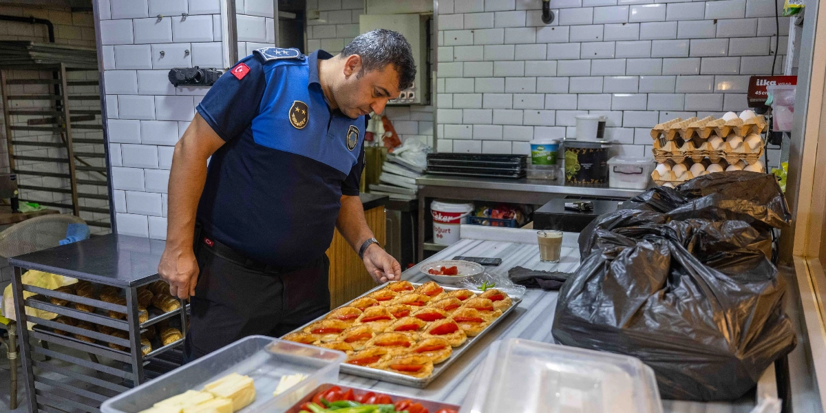 Mavi ve lacivert üniformalı bir zabıta memuru, beyaz fayanslı bir mutfak tezgahının üzerinde duran domatesli hamur işlerini inceliyor. Tezgahın üzerinde ayrıca büyük siyah çöp poşetleri, bir bardak çay ve arka planda yumurta kolileri bulunuyor.