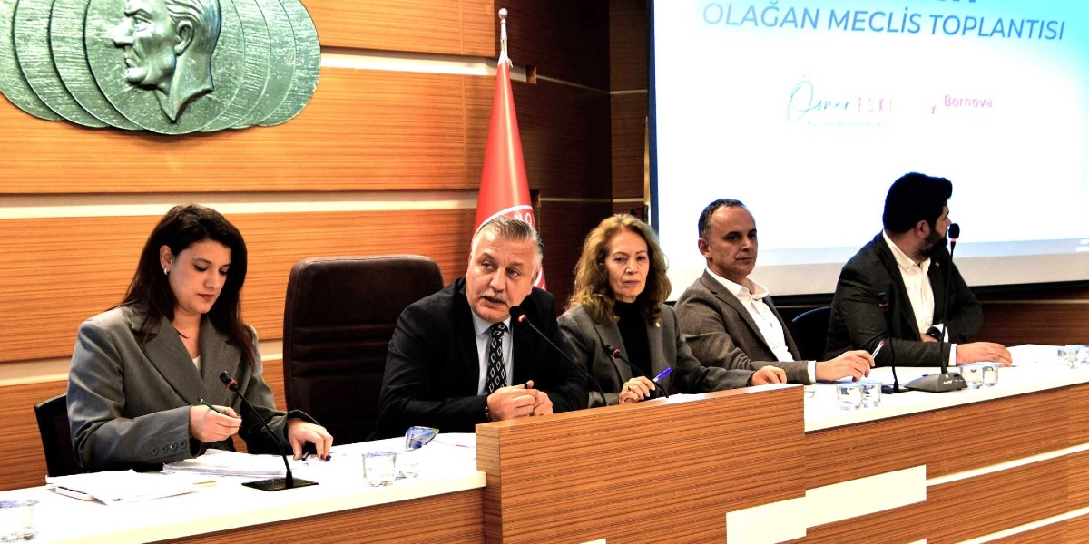 Bornova Belediyesi meclis toplantısında, masada yan yana oturan beş kişilik bir heyet yer alıyor. En solda Gülşah Durbay, yanında ise Bornova Belediye Başkanı Ömer Eşki ve diğer meclis üyeleri bulunuyor. Heyetin arkasında ahşap kaplamalı duvarda Atatürk kabartması ve Türk bayrağı yer alırken, sağ tarafta üzerinde 