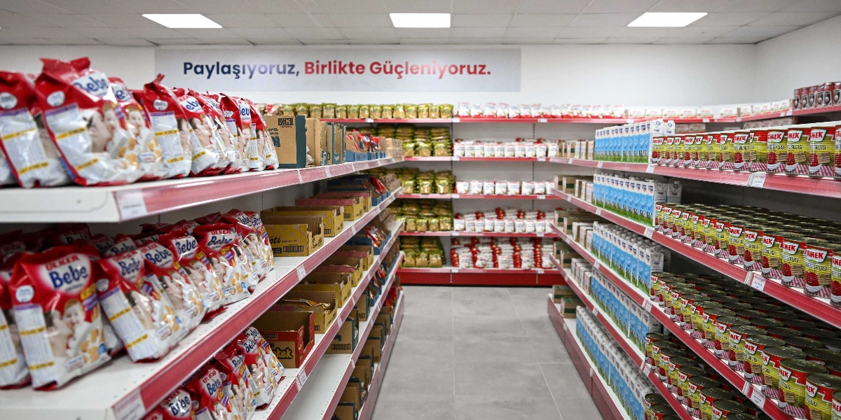 Bir market reyonunda raflar üzerinde düzenli bir şekilde dizilmiş bebek bisküvileri, süt kutuları ve konserve gıdalar görülüyor. Arka duvarda 