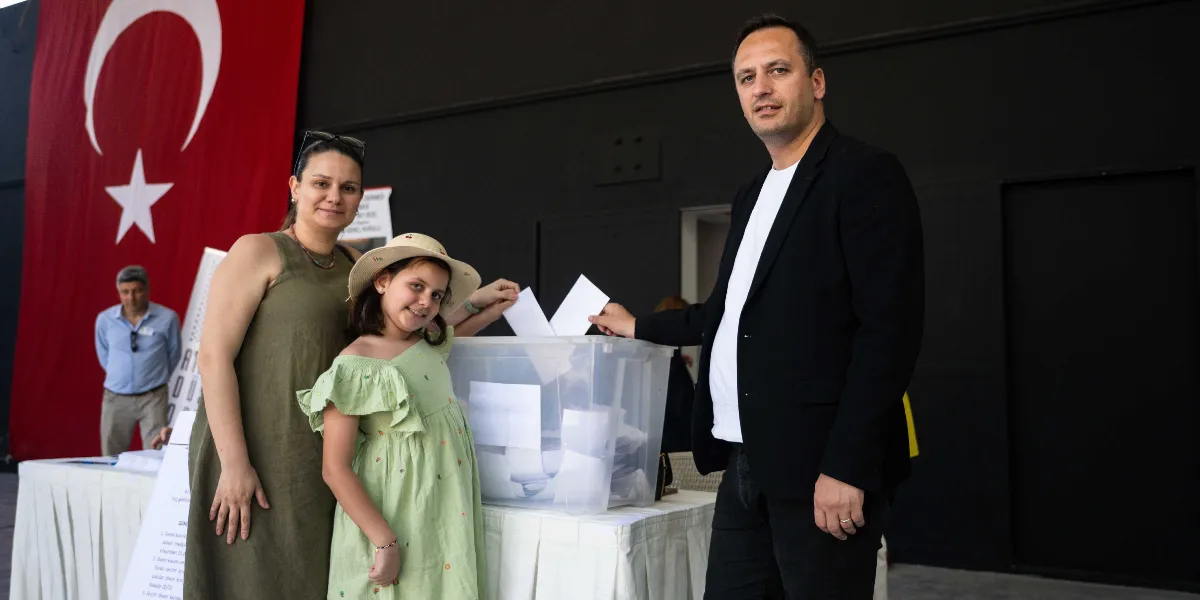 Atatürkçü Düşünce Derneği Genel Kurulu'nda, siyah ceketli bir erkek, yanında yeşil elbiseli bir kız çocuğu ve zeytin yeşili elbiseli bir kadınla birlikte şeffaf bir seçim sandığına oy pusulası atıyor. Arka planda büyük bir Türk bayrağı ve masanın başında duran bir görevli yer alıyor.