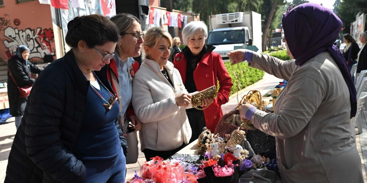 Açık havada kurulan bir stantta, el yapımı takı ve hediyelik eşyaları inceleyen dört kadın ve onlara ürünleri gösteren başörtülü bir satıcı kadın yer alıyor. Kadınlar, masanın üzerindeki çantaları ve çiçekli süslemeleri dikkatle inceliyor. Arka planda ise duvarında grafiti bulunan bir bina, park halindeki bir kamyonet ve yürüyüş yolunda duran diğer insanlar görülüyor.