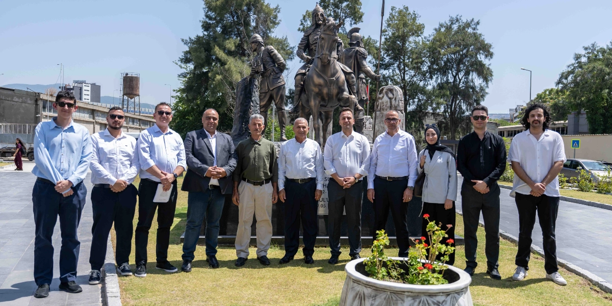 Açık havada, arkalarında atlı ve asker figürlerinden oluşan bronz bir anıtın önünde yan yana duran, çoğunluğu erkeklerden oluşan on kişilik bir grup. Gruptaki kişiler genellikle açık renkli gömlekler ve koyu renkli pantolonlar giymiş, bazıları güneş gözlüğü takmış durumdadır. Grubun önünde, içinde çiçekler bulunan beyaz bir saksı yer almaktadır. Arka planda ağaçlar, bir yol ve binalar görülmektedir.