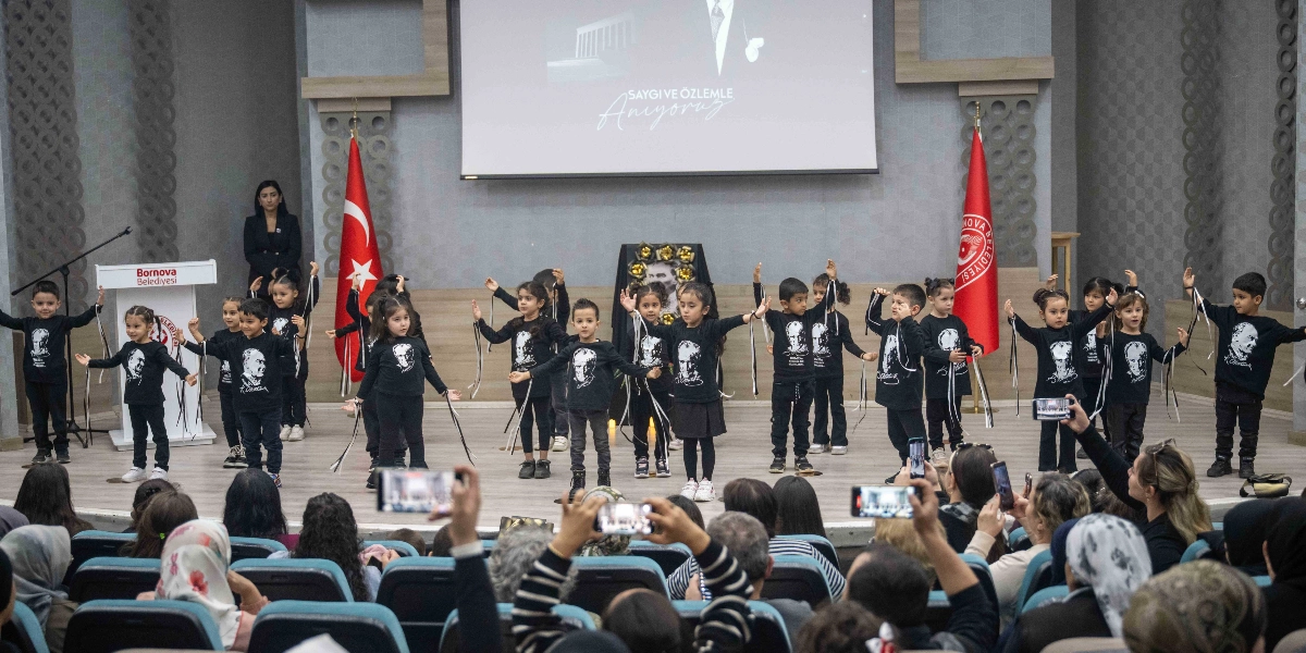Üzerinde Atatürk silüeti bulunan siyah tişörtler giymiş bir grup çocuk, bir sahnede ellerinde siyah beyaz kurdelelerle gösteri yapıyor. Sahnenin arka planındaki dev ekranda 