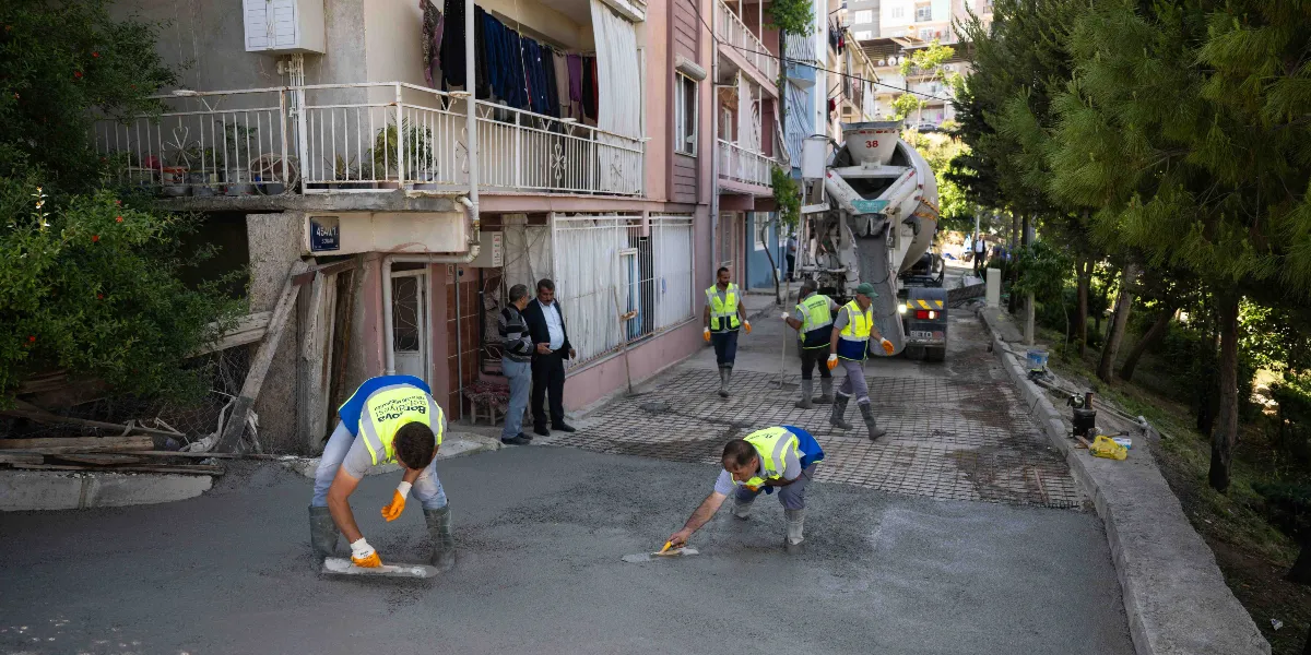 Bornova Belediyesi ekipleri, yokuşlu bir sokakta beton dökme ve düzeltme çalışması yapıyor. Sarı yelekli işçiler ellerindeki mala ve ekipmanlarla taze betonu düzeltirken, arka planda bir beton mikseri ve çalışmayı izleyen iki kişi bulunuyor.