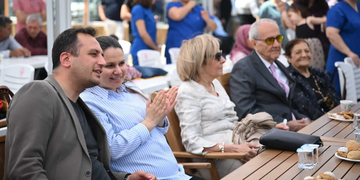 Açık havada düzenlenen bir etkinlikte, yan yana oturan bir grup insan bir konuşmayı dinleyip alkışlıyor. Ön planda, gri ceketli bir erkek ve yanında mavi beyaz çizgili gömlek giymiş, elleriyle alkışlayan bir kadın bulunuyor. Onların hemen yanında, güneş gözlüklü, açık renkli kıyafetli bir kadın, takım elbiseli ve sarı camlı gözlük takan yaşlı bir beyefendi ile koyu renkli kıyafetli gülen bir kadın oturuyor. Masanın üzerinde tabaklarda kurabiyeler ve su bardakları yer alıyor. Arka planda ise ayakta duran ve oturan diğer katılımcılar görülüyor.