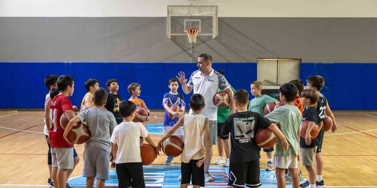 Kapalı bir spor salonunda, basketbol antrenörü eşliğinde çember oluşturmuş bir grup çocuk basketbol antrenmanı yapıyor. Antrenör, elinde bir basketbol topu tutarak çocuklara talimat veriyor; çocuklar ise ellerinde toplarla onu dikkatle dinliyor. Salonun zemininde Bornova Belediyesi logosu yer alıyor.