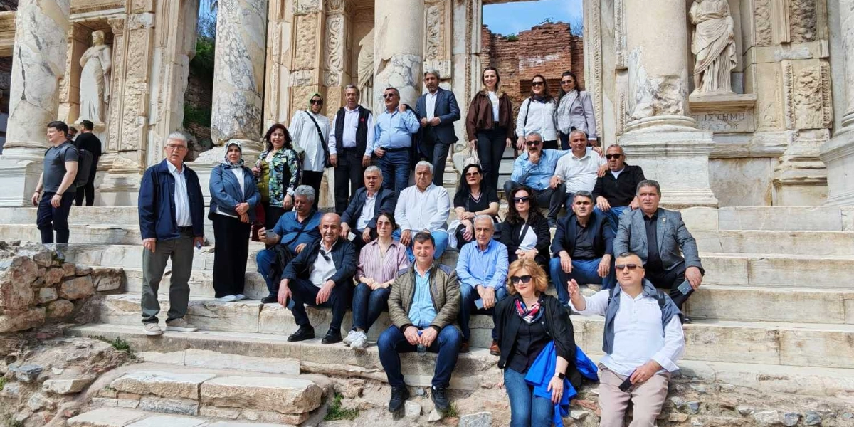 Efes Antik Kenti'ndeki Celsus Kütüphanesi'nin önündeki basamaklarda poz veren, Bornova Muhtarlar Akademisi katılımcılarından oluşan kalabalık bir grup. Kadın ve erkeklerden oluşan grup üyeleri, tarihi sütunlar ve heykellerin önünde yan yana ve oturur vaziyette durarak toplu bir hatıra fotoğrafı çektiriyor.