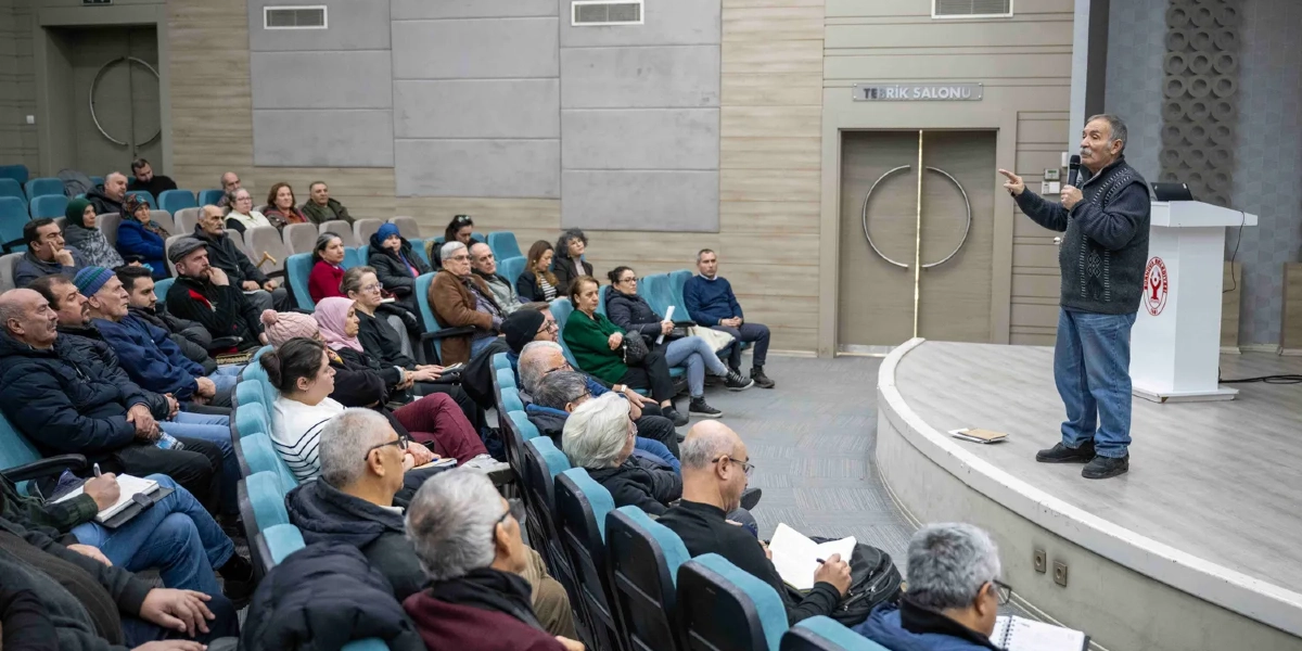 Modern bir konferans salonunda, kürsüde elinde mikrofonla konuşma yapan yaşlı bir adam ve onu dikkatle dinleyen, çoğunluğu orta yaş ve üzeri kişilerden oluşan bir izleyici grubu. Salonun duvarında 