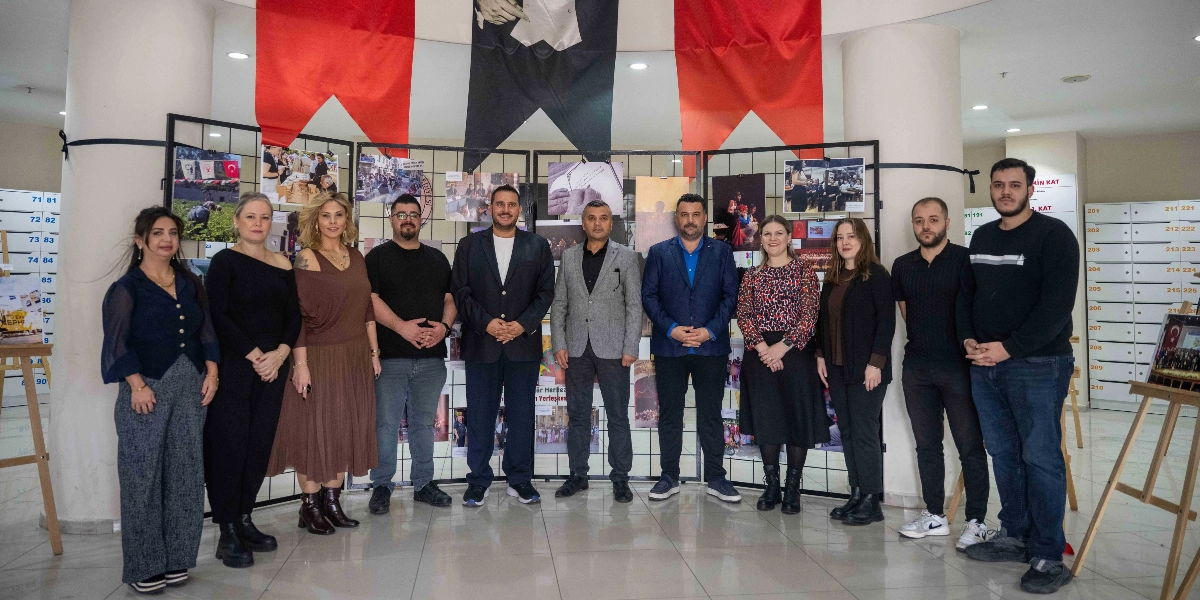 Bornova'da düzenlenen Birlikte Mümkün sergisinde, arka planda çeşitli fotoğrafların asılı olduğu bir sergi panosunun önünde yan yana duran on bir kişilik bir grup. Grubun arkasında, ortada Atatürk portresi ve iki yanında kırmızı bayraklar yer alıyor. Katılımcılar, günlük ve şık kıyafetler içinde, gülümseyerek kameraya bakıyor. Mekan, beyaz sütunları ve posta kutularını andıran bölmeleriyle aydınlık bir iç mekan görünümünde.