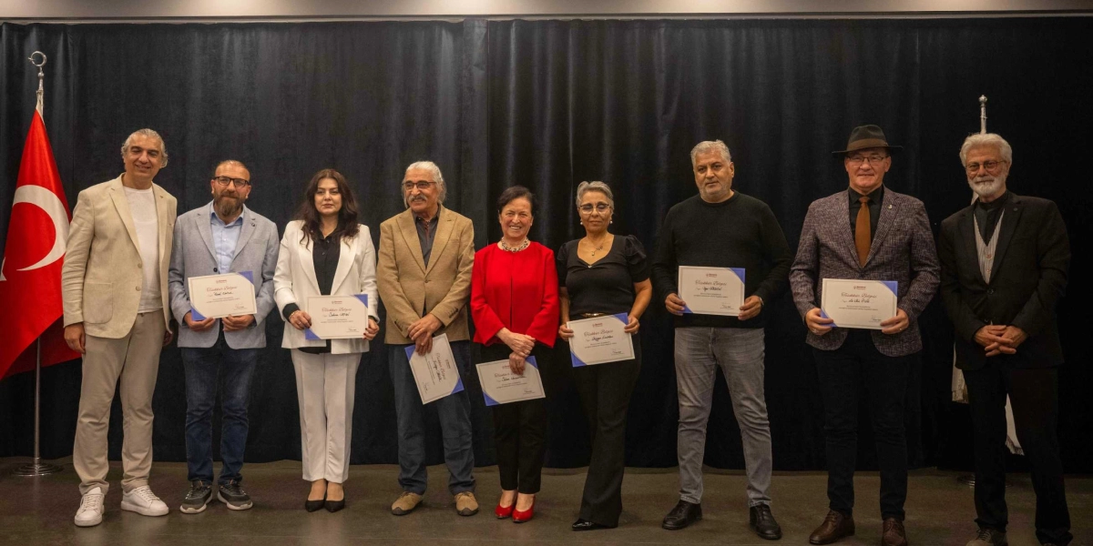 Koyu renkli bir perde önünde yan yana duran dokuz kişilik grup, ellerinde Bornova Belediyesi logolu teşekkür belgeleriyle poz veriyor. Solda bir Türk bayrağı, sağda ise bir direk yer alıyor. Gruptaki kişiler, günlük ve yarı resmi kıyafetler içinde, gülümseyerek kameraya bakıyor.
