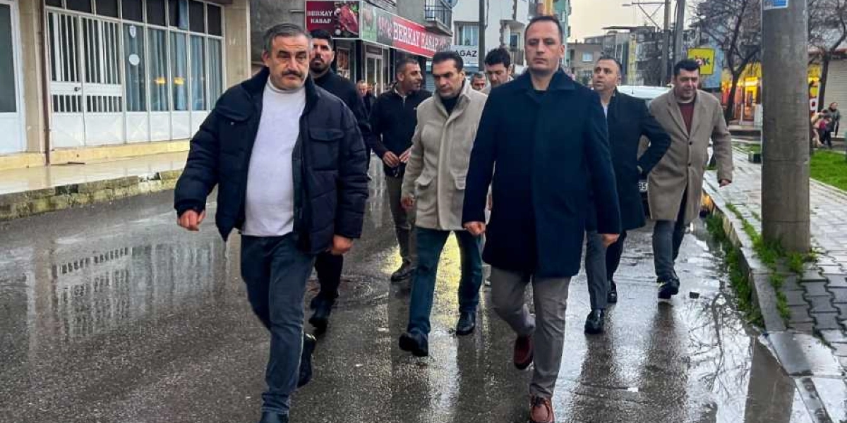 Kışlık kıyafetler giymiş bir grup erkek, ıslak bir sokakta yan yana yürüyerek ilerliyor. Grubun önünde koyu renk montlu ve bej kabanlı kişiler yer alırken, arkalarında onları takip eden diğer kişilerle birlikte bir inceleme gezisi izlenimi veriyorlar. Arka planda dükkanların bulunduğu bir cadde ve yer yer su birikintileri olan asfalt bir yol görülüyor.