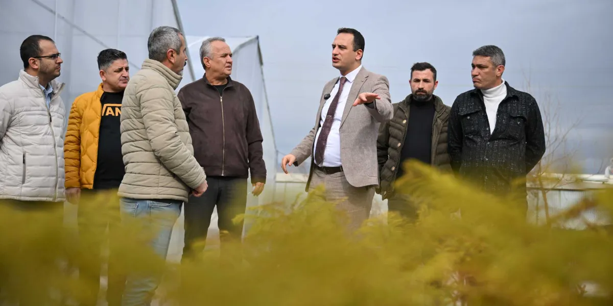 Modern bir seranın içinde, takım elbiseli bir adamın çevresindeki beş kişilik gruba bir şeyler anlattığı ve eliyle işaret ettiği bir toplantı anı. Gruptaki diğer kişiler günlük kıyafetler içinde, ilgiyle konuşmacıyı dinliyor. Ön planda bulanık bir şekilde görünen yeşil bitkiler, arka planda ise seranın plastik kaplamaları yer alıyor.
