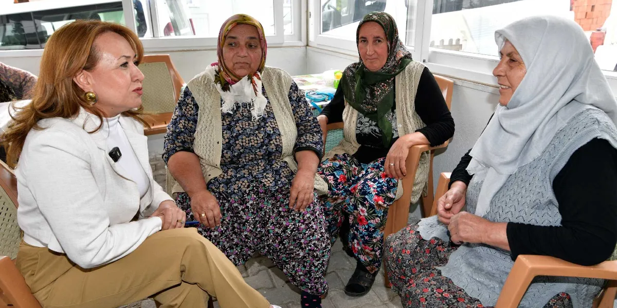 Açık renkli ceket giymiş bir kadın, köy ortamında oturan üç kadınla sohbet ederek sağlık eğitimi veriyor. Kadınlar plastik sandalyelerde oturuyor ve geleneksel kıyafetler ile başörtüsü kullanıyorlar.