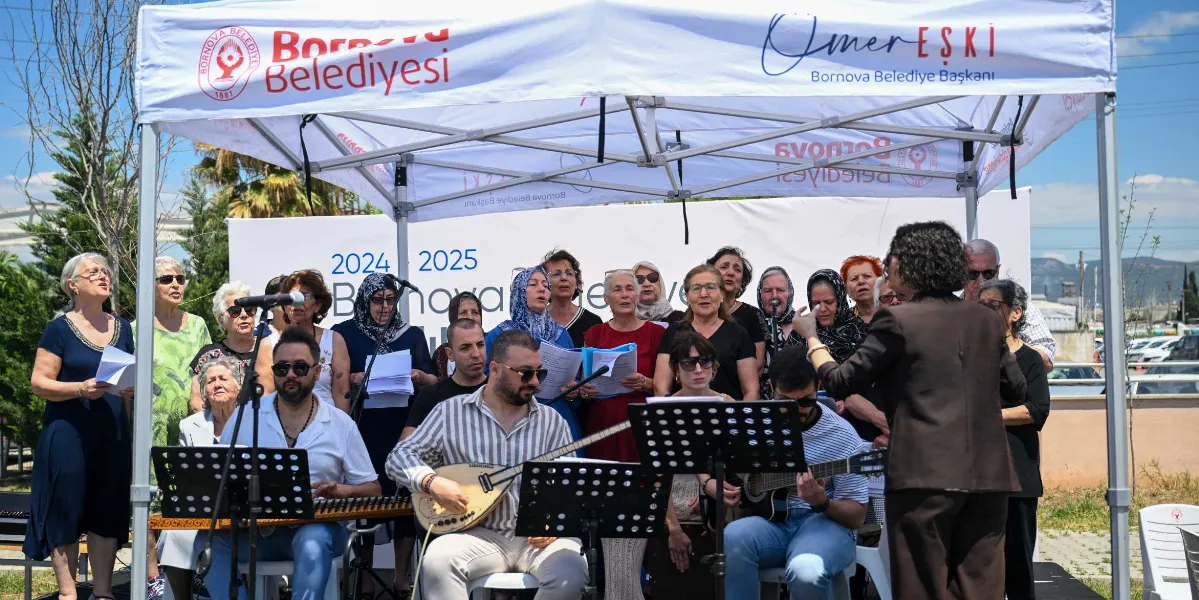 Açık havada, Bornova Belediyesi logolu beyaz bir çadırın altında toplanmış bir koro ve müzisyenler grubu. Ön planda saz çalan bir erkek, gitar çalan bir genç ve onları yöneten bir şef bulunuyor. Arkalarında ellerinde notalarla şarkı söyleyen, çoğunluğu orta yaş ve üzeri kadınlardan oluşan bir koro yer alıyor. Arka plandaki panoda 2024-2025 dönemiyle ilgili ifadeler görülüyor.