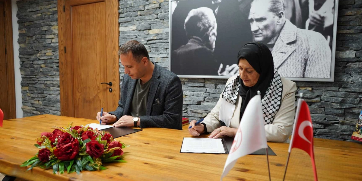 Bornova Belediye Başkanı ve Filistin'in Dura Belediyesi'nden bir temsilci, ahşap bir masa başında kardeş şehir protokolünü imzalıyor. Masanın üzerinde kırmızı çiçeklerden oluşan bir aranjman ile Türk ve Filistin bayrakları yer alıyor. Arka planda taş duvar üzerinde Mustafa Kemal Atatürk'ün siyah beyaz bir portresi bulunuyor.