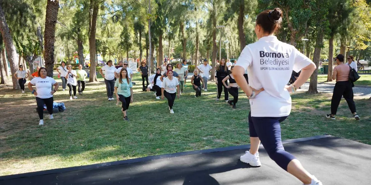 Ağaçlık bir park alanında, Bornova Belediyesi Spor İşleri Müdürlüğü tişörtü giyen bir eğitmen eşliğinde egzersiz yapan bir grup kadın. Eğitmen önde, siyah bir platform üzerinde durarak katılımcılara hareketleri gösterirken, katılımcılar çimenlik alanda aynı hareketleri tekrarlıyor.