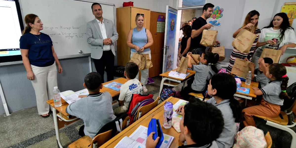 Bir sınıf ortamında, Bornova Belediyesi logolu kahverengi kağıt çantalar içindeki beslenme paketlerini öğrencilere dağıtan belediye yetkilileri ve gönüllüler görülüyor. Sınıfın önünde beyaz tahtanın yanında duran bir kadın öğretmen ve gri ceketli bir erkek yetkili öğrencilere eşlik ederken, diğer yetişkinler sıraların arasında dolaşarak öğrencilere paketlerini veriyor. Öğrenciler sıralarında oturmuş, ders kitapları ve kırtasiye malzemeleriyle birlikte kendilerine sunulan desteği alıyorlar.