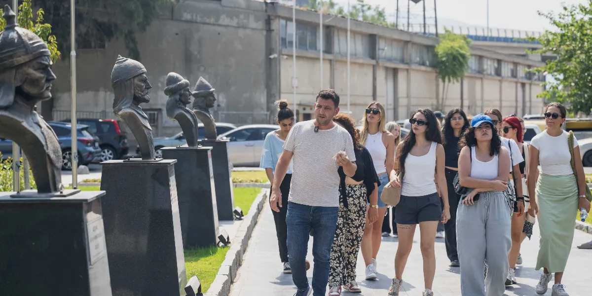 Abide-i Hürriyet Meydanı’nda, bir rehber eşliğinde yürüyen gençlerden oluşan bir grup, yol kenarındaki tarihi büstleri inceliyor. Grubun sol tarafında, kaideler üzerinde sıralanmış Osmanlı dönemi figürlerini temsil eden bronz büstler yer alırken, sağ tarafta ise günlük kıyafetleri içindeki ziyaretçiler güneşli bir havada yürüyüşlerine devam ediyor. Arka planda ise beton bir duvar ve otopark alanı görülüyor.