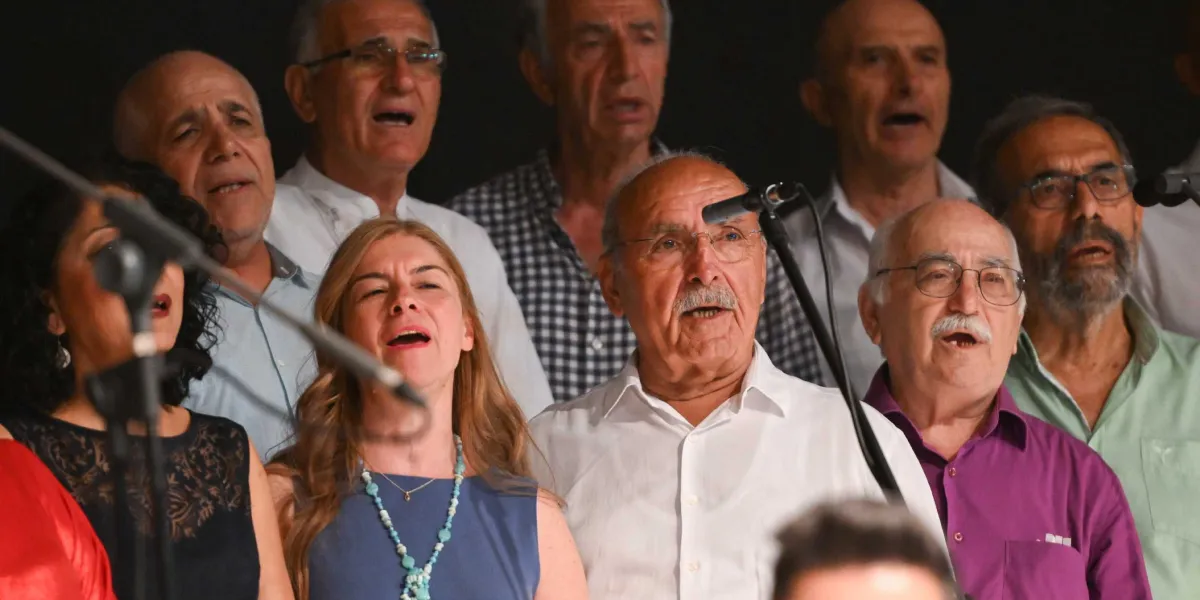Sahne üzerinde şarkı söyleyen, farklı yaş gruplarından oluşan bir koro üyeleri. Ön planda mavi elbiseli ve turkuaz kolyeli bir kadın ile beyaz gömlekli, gözlüklü ve bıyıklı bir erkek yer alıyor. Arka planda ise çeşitli kıyafetler giymiş, ağızları açık şekilde şarkı söylemeye devam eden diğer koro üyeleri bulunuyor. Önlerinde mikrofon standları yer alıyor ve ortamın arka planı karanlık.