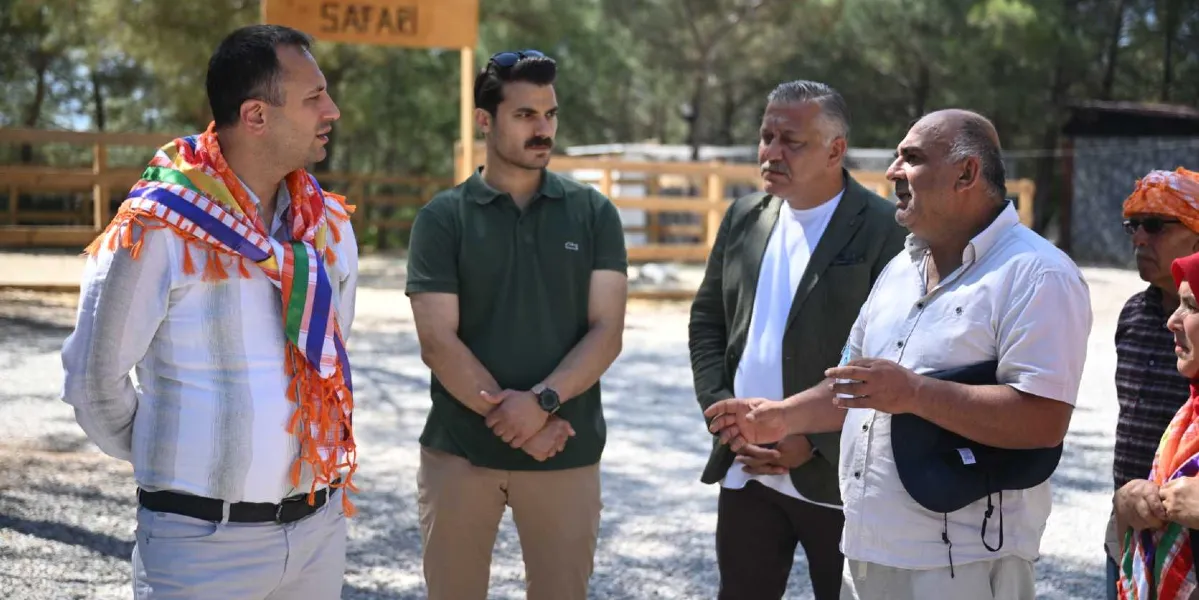 Açık havada, ağaçlık bir alanda bir araya gelen bir grup insan, birbirleriyle sohbet ediyor. En önde, beyaz gömlekli ve elinde lacivert bir şapka tutan orta yaşlı bir adam konuşurken, karşısındaki diğer kişiler onu dinliyor. Dinleyenler arasında omuzlarında renkli bir şal bulunan beyaz gömlekli bir erkek, haki renkli polo yaka tişört giyen genç bir erkek ve ceketli bir başka erkek yer alıyor. Arka planda ahşap çitler ve üzerinde 