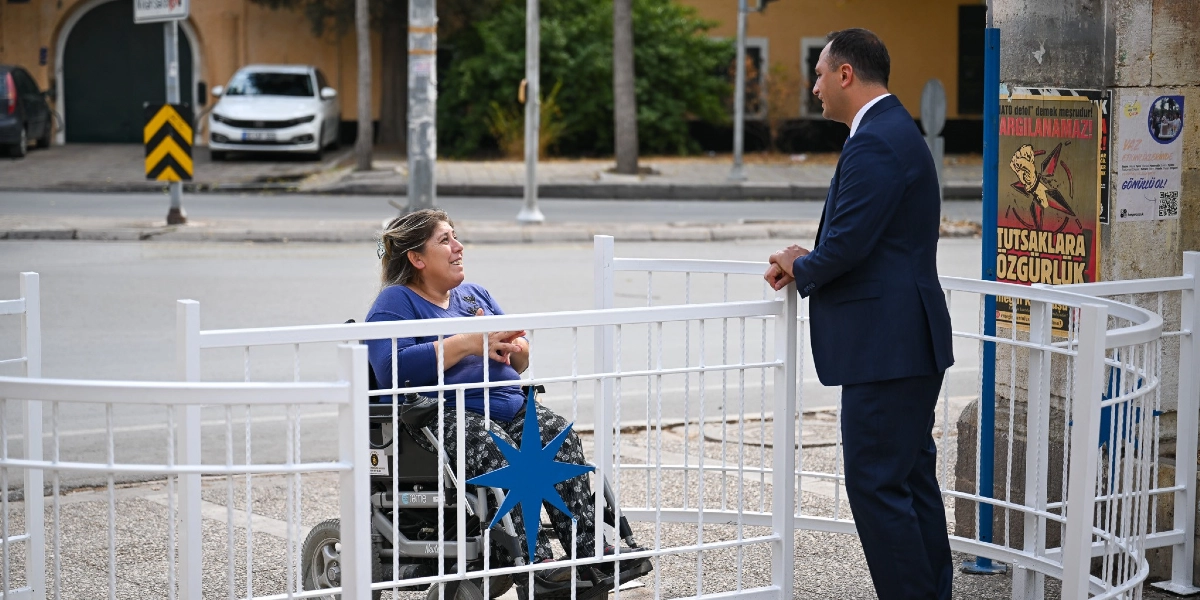 Tekerlekli sandalyede oturan bir kadın, Bornova Belediyesi tarafından park girişlerine yerleştirilen yeni nesil bariyerlerin önünde, takım elbiseli bir yetkiliyle sohbet ediyor. Beyaz metalden yapılmış ve üzerinde mavi yıldız figürü bulunan bariyerler, motosiklet girişini engellerken tekerlekli sandalye kullanımına uygun bir geçiş alanı sağlıyor.