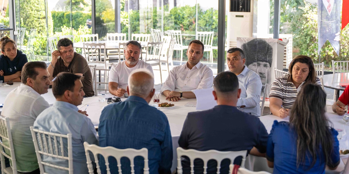 Aydınlık ve camlarla çevrili bir mekânda, yuvarlak bir masa etrafında toplanmış bir grup insan toplantı yapıyor. Masanın başında Bornova Belediye Başkanı Ömer Eşmi'nin de aralarında bulunduğu katılımcılar, önlerindeki belgeleri inceliyor ve birbirleriyle iletişim kuruyor. Masanın arka planında Mustafa Kemal Atatürk'ün bir portresinin yer aldığı bir pano bulunuyor. Katılımcılar gündelik kıyafetler içinde, masada çay bardakları ve notlar yer alıyor.