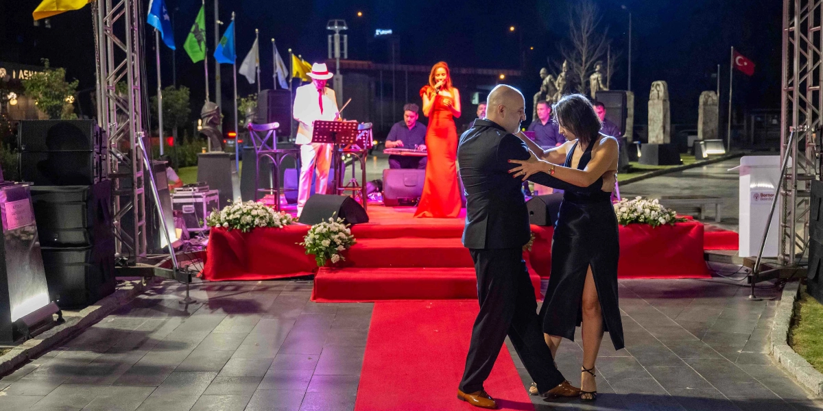 Gece vakti açık havada düzenlenen bir etkinlikte, kırmızı halı üzerinde tango yapan bir çift ve arkalarındaki sahnede canlı müzik yapan bir grup görülüyor. Sahnede beyaz takım elbiseli ve şapkalı bir müzisyen, kırmızı uzun elbiseli bir kadın solist ve enstrüman çalan diğer grup üyeleri yer alıyor. Arka planda ise direklerde asılı bayraklar, heykeller ve Bornova Belediyesi'ne ait bir kürsü bulunuyor.