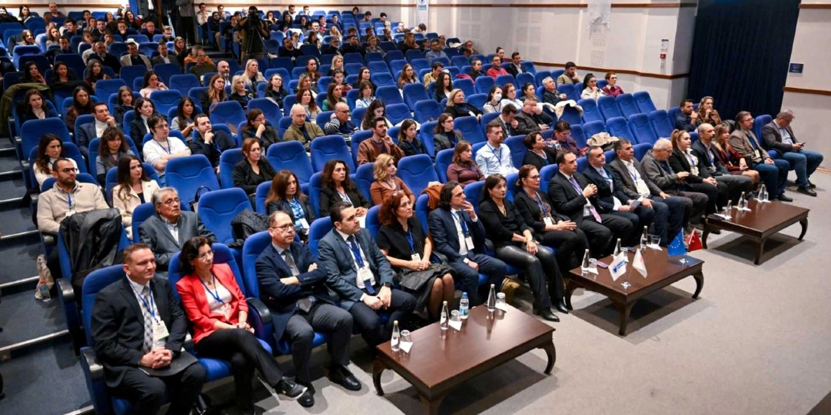 Konferans salonunda mavi koltuklarda oturan kalabalık bir dinleyici grubu, sahneye dönük şekilde oturuyor. Ön sıralarda takım elbiseli ve resmi kıyafetli katılımcılar yer alırken, salonun genelinde farklı yaş gruplarından insanlar dikkatle dinliyor. Ön planda, üzerinde küçük bayraklar ve isimlikler bulunan ahşap sehpalar yer alıyor. Salonun genel atmosferi resmi bir toplantı veya semineri yansıtıyor.