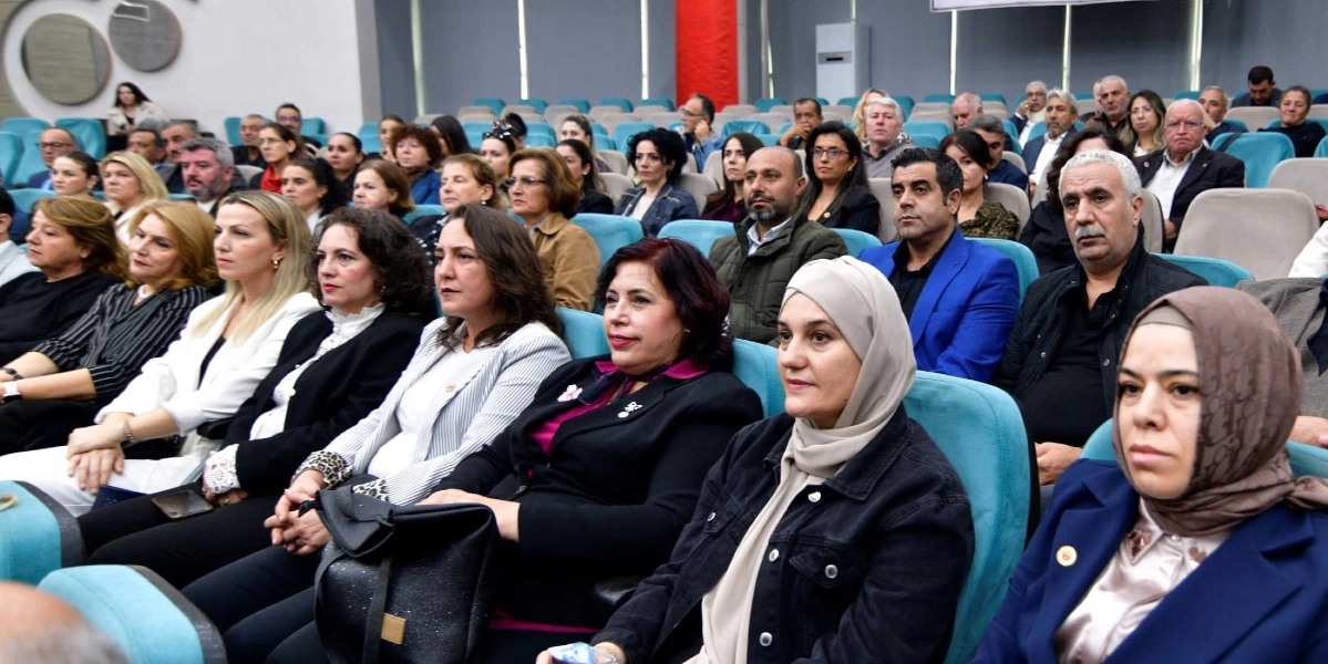 Konferans salonunda yan yana dizilmiş turkuaz renkli koltuklarda oturan, farklı yaş ve giyim tarzlarına sahip kalabalık bir grup muhtar adayı ve katılımcı, dikkatle karşıya bakarak bir sunumu dinliyor. Ön planda, başörtülü kadınlar, ceket giymiş erkekler ve çeşitli kıyafetlerle oturan kadınlar yer alıyor; arka sıralara doğru katılımcı sayısı artıyor. Salonun genelinde resmi ve ciddi bir atmosfer hakim.