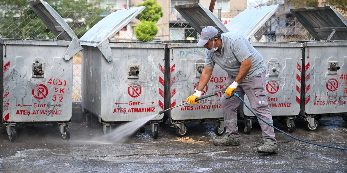 Belediye üniformalı, şapkalı ve maskeli bir görevli, yan yana dizilmiş metal çöp konteynerlerinin bulunduğu alanda basınçlı su tabancasıyla zemin temizliği yapıyor. Konteynerlerin üzerinde 