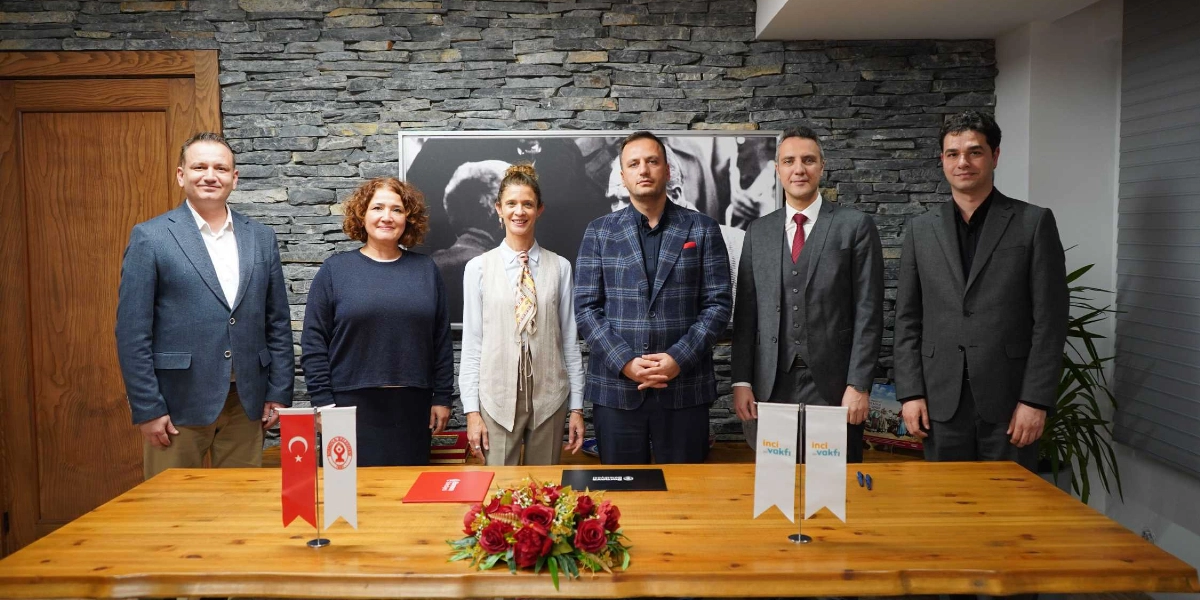 Taş duvarlı bir odada, ahşap bir masanın arkasında yan yana duran altı kişi, iş birliği protokolü imza töreni için poz veriyor. Masanın üzerinde Türk bayrağı, Bornova Belediyesi logosu ve İnci Vakfı logolu küçük bayraklar ile bir çiçek aranjmanı bulunuyor. Katılımcılar, resmi kıyafetler içinde gülümseyerek kameraya bakıyor; arka planda ise siyah beyaz bir Atatürk portresi yer alıyor.