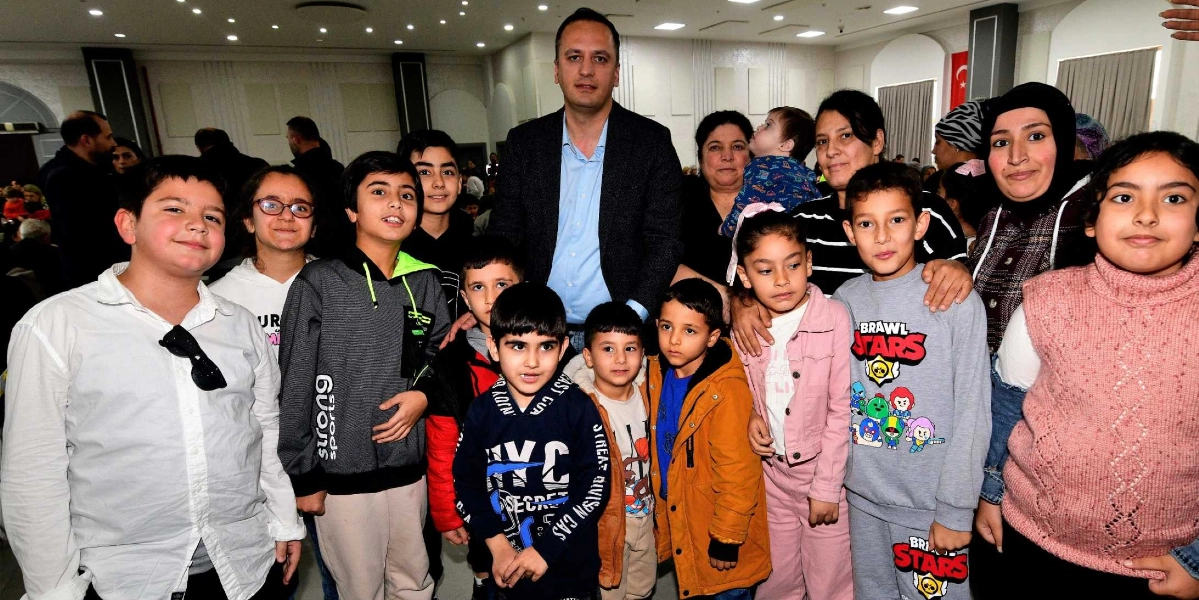 Bornova Belediye Başkanı Ömer Eşki, geniş bir salonda düzenlenen yılbaşı yemeğinde çocuklarla ve aileleriyle bir araya gelerek hatıra fotoğrafı çektiriyor. Başkan Eşki, koyu renkli ceketi ve açık mavi gömleğiyle grubun merkezinde yer alırken, etrafında farklı yaş gruplarından çocuklar ve bazı yetişkinler gülümsüyor. Çocuklardan bazıları Brawl Stars baskılı kıyafetler giyiyor. Arka planda ise yemek salonunun aydınlatmaları ve diğer davetliler seçiliyor.