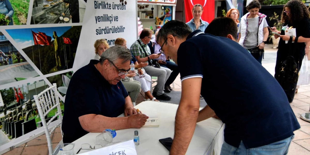 İlber Ortaylı, Bornova'da düzenlenen bir etkinlikte masasında oturmuş, hayranı için kitabını imzalıyor. Ortaylı'nın karşısında duran bir kişi, imza sürecini izlerken masanın arkasında ise etkinliği takip eden diğer katılımcılar yer alıyor. Arka planda üzerinde 