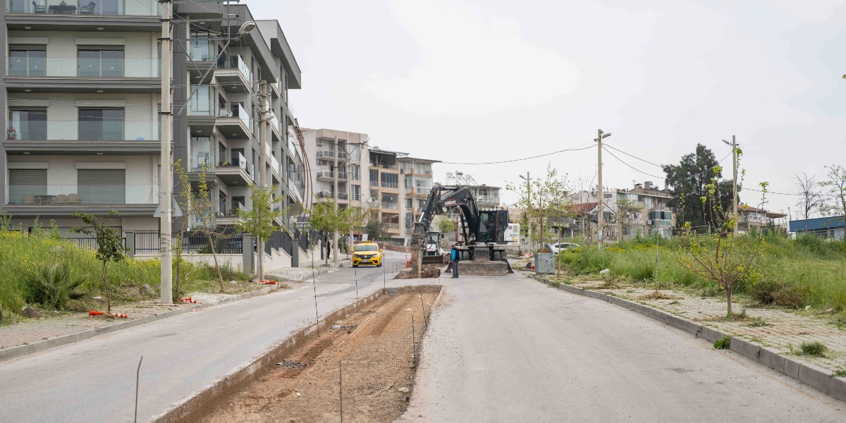 Bornova'da bir yol çalışması alanında, asfaltı kıran bir iş makinesi ve başında duran bir operatör görülüyor. Yolun ortası kazılmış durumda ve arka planda modern apartmanlar ile sarı bir ticari taksi yer alıyor.