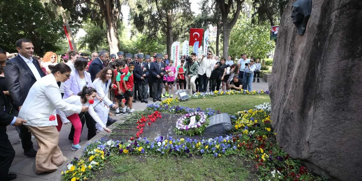 Açık havada, ağaçlık bir alanda bulunan Zübeyde Hanım büstünün ve anıtının önünde düzenlenen Anneler Günü anma töreni. Beyaz ceketli bir kadın ve beraberindeki genç kız, anıtın önüne kırmızı karanfiller bırakıyor. Arka planda Türk bayrağı, resmi kıyafetli kişiler, çocuklar ve töreni takip eden kalabalık bir grup yer alıyor. Anıtın çevresi renkli çiçeklerle süslenmiş durumda.