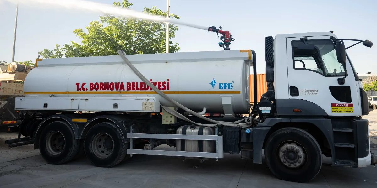 Üzerinde T.C. Bornova Belediyesi yazılı beyaz bir itfaiye veya su tankeri aracı, tepe kısmındaki mekanizmadan havaya doğru tazyikli su püskürtüyor. Aracın kapısında Bornova Belediyesi logosu ve şoförün görüş açısıyla ilgili uyarı yazısı bulunuyor.