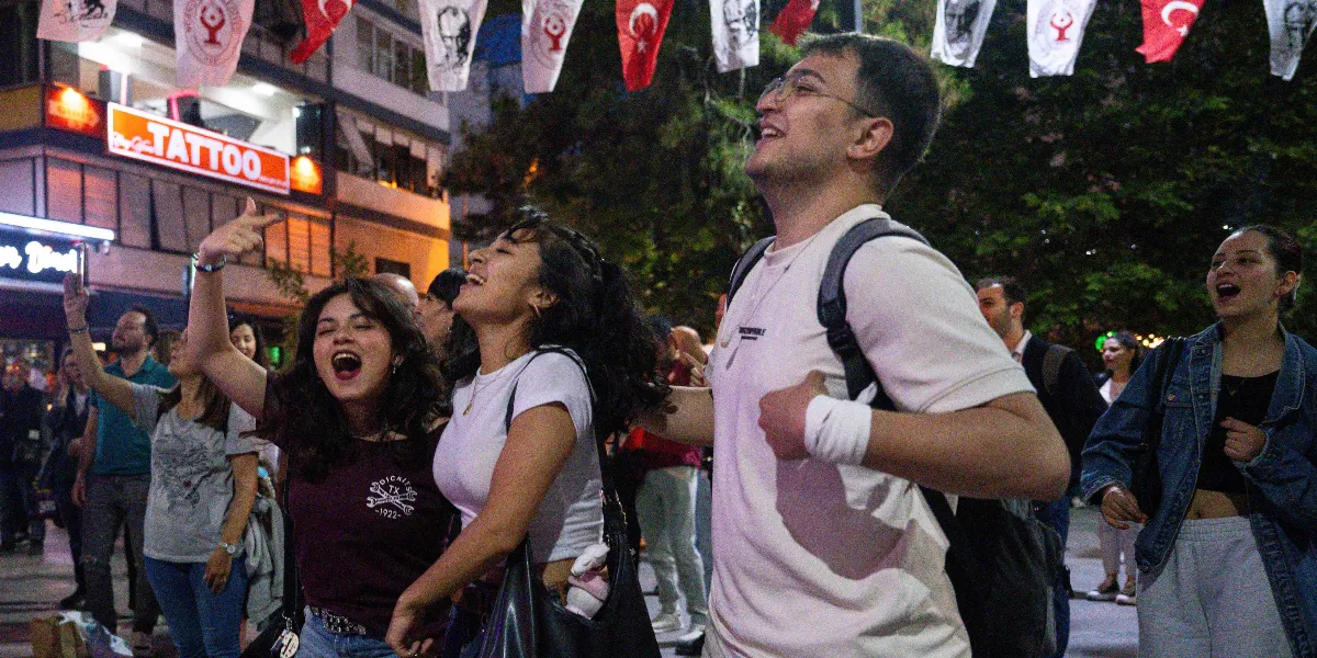 Gece vakti bir sokak kutlamasında, ellerini havaya kaldırarak neşeyle şarkı söyleyen ve dans eden genç bir grup insan. Arka planda Bornova'daki dükkanların ışıklı tabelaları ve yukarıda asılı duran Türk bayrakları ile Atatürk silüetli flamalar görülüyor.
