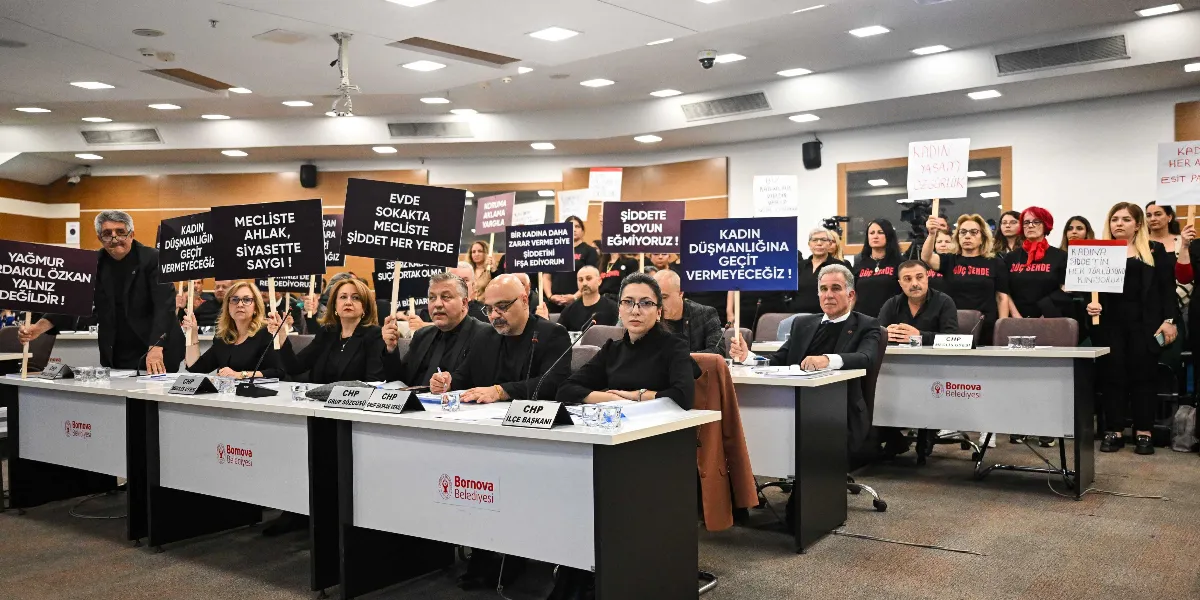 Bornova Belediyesi meclis salonunda, siyah giyinmiş bir grup kadın ve erkek, kadına yönelik şiddeti protesto etmek amacıyla ellerinde pankartlarla oturuyor ve ayakta duruyor. Pankartlarda 