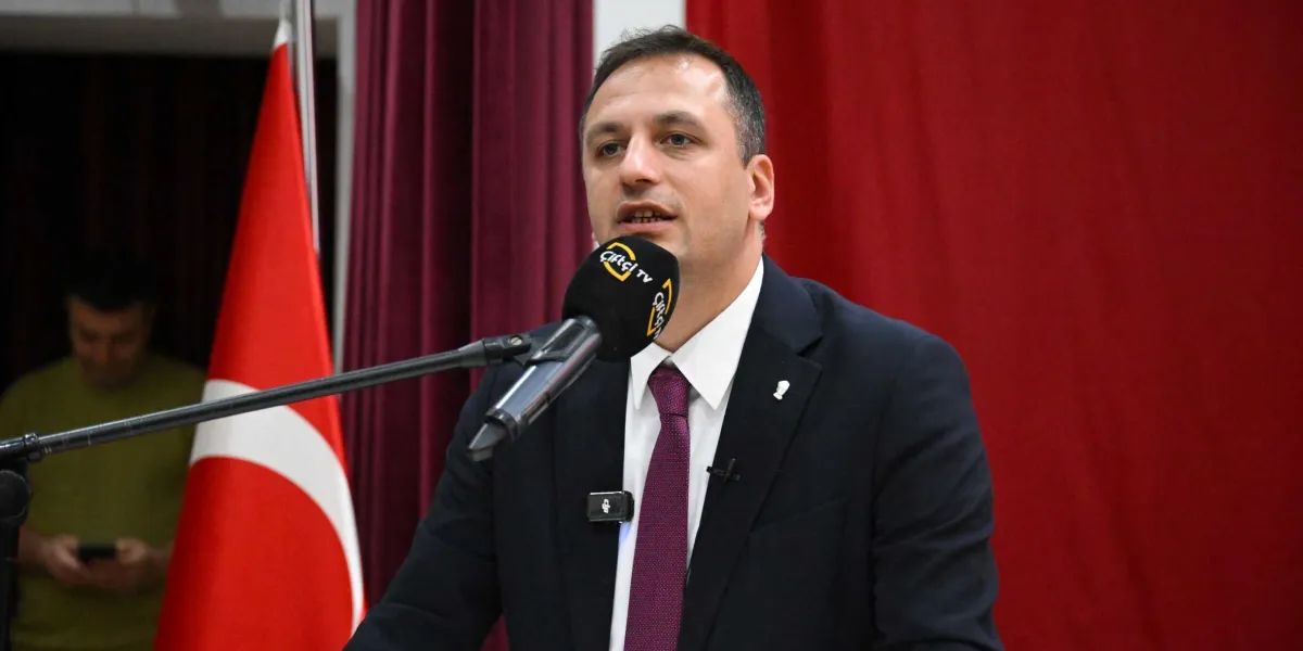 Koyu renk takım elbise ve bordo kravat giymiş bir erkek, önündeki mikrofonla konuşma yapıyor. Arka planda kırmızı bir perde ve Türk bayrağı yer alıyor. Mikrofonun üzerinde Çiftçi TV logosu bulunuyor.