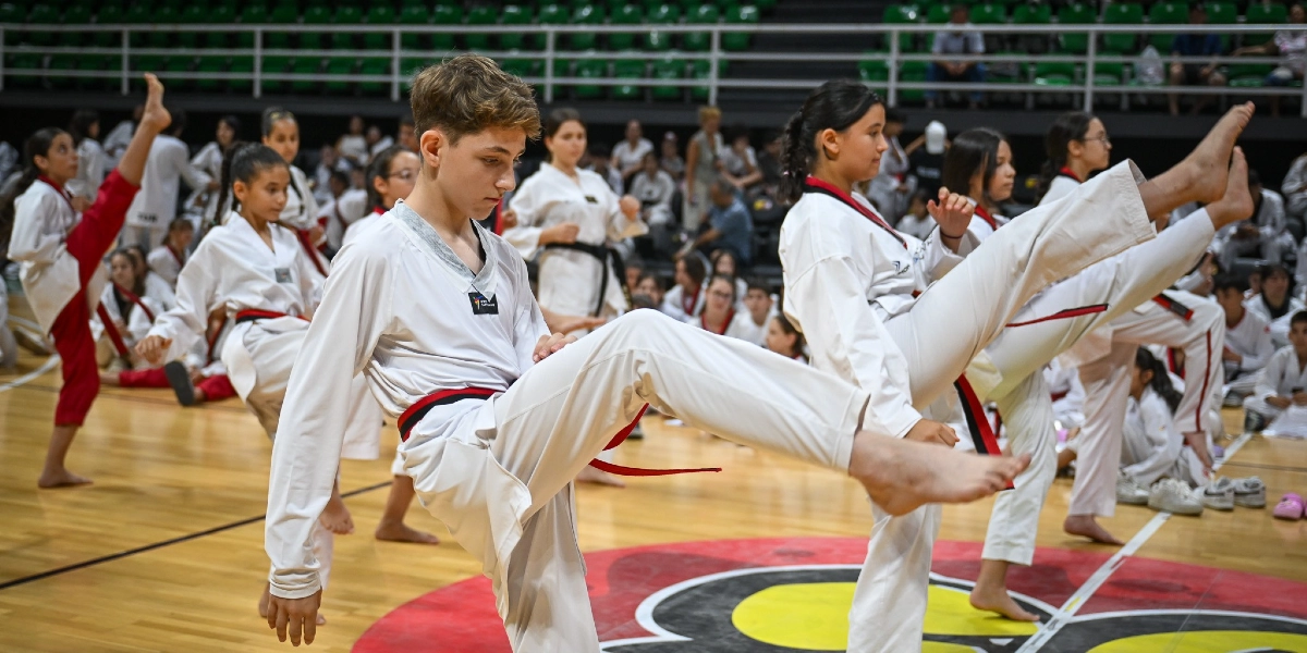 Spor salonunda beyaz tekvando kıyafetleri giymiş bir grup genç, eğitmenleri eşliğinde senkronize bir şekilde tekme atma hareketi çalışıyor. Ön planda odaklanmış bir erkek öğrenci ve yanında benzer hareketi yapan kız öğrenciler bulunuyor. Arka planda ise tribünlerde oturan izleyiciler ve diğer sporcular yer alıyor.