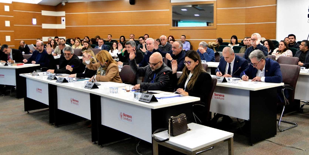 Bornova Belediyesi meclis salonunda düzenlenen toplantıda, masalarda oturan çok sayıda katılımcı ellerini kaldırarak oylamaya katılıyor. Ön sırada, üzerinde 