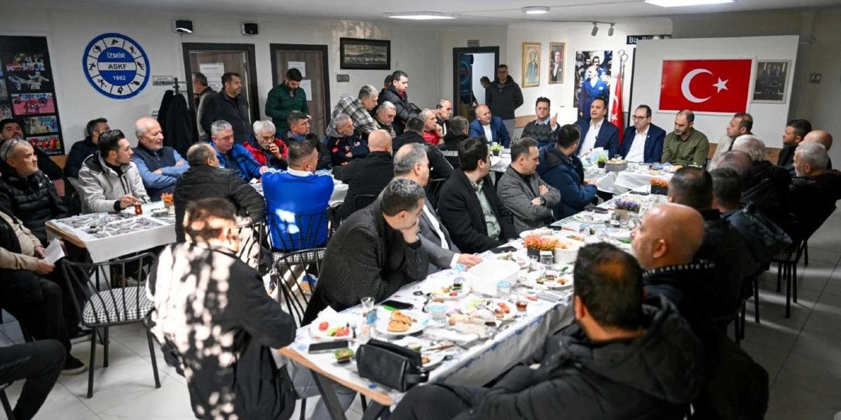 İzmir Amatör Spor Kulüpleri Federasyonu binasında, uzun masalar etrafında toplanmış kalabalık bir grup erkek, yemekli bir toplantı sırasında bir araya gelmiş durumda. Masalarda tabaklar, çay bardakları ve çeşitli ikramlar bulunurken, katılımcılar dikkatle konuşmacıları dinliyor. Duvarlarda İzmir ASKF logosu, Atatürk portreleri ve Türk bayrağı asılı. Ortamda resmi bir toplantı havası hakim.