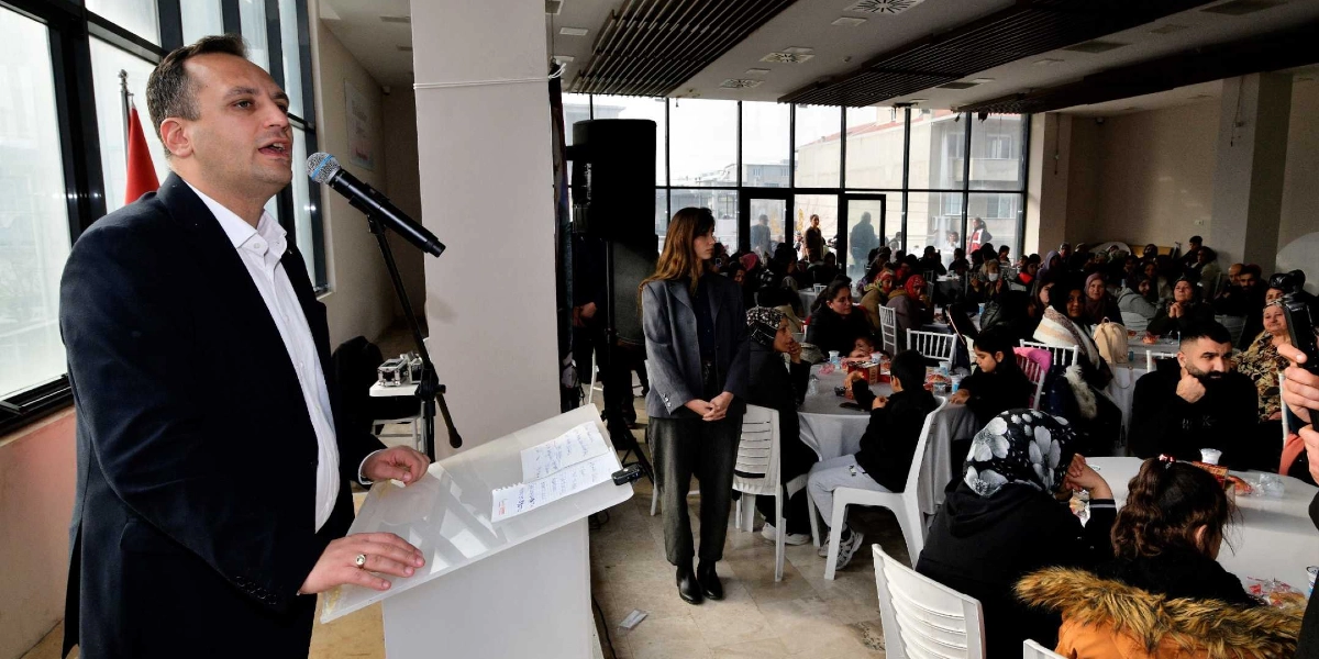 Bornova Belediye Başkanı Ömer Eşki, kürsüde mikrofon başında konuşma yaparken, arka planda yılbaşı yemeğine katılan çok sayıda Bornovalı vatandaş masalarda oturuyor.