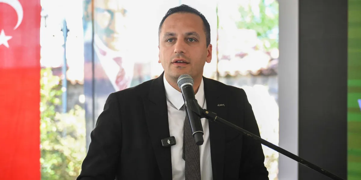 Siyah takım elbiseli, beyaz gömlekli ve gri kravatlı bir erkek, mikrofon başında konuşma yapıyor. Arka planda kısmen görünen bir Türk bayrağı ve bulanık bir Atatürk portresi yer alıyor.