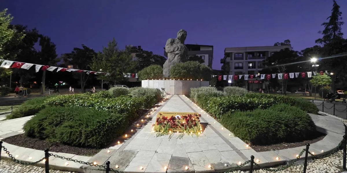 Alacakaranlıkta, Bornova'daki Srebrenica Soykırım Anıtı'nın önünde, üzerinde çiçekler bulunan bir kaide ve anıtın çevresine dizilmiş çok sayıda küçük mum yer alıyor. Anıtın etrafındaki ağaçlar arasına asılmış Türk bayrakları ve anma flamaları görülüyor.