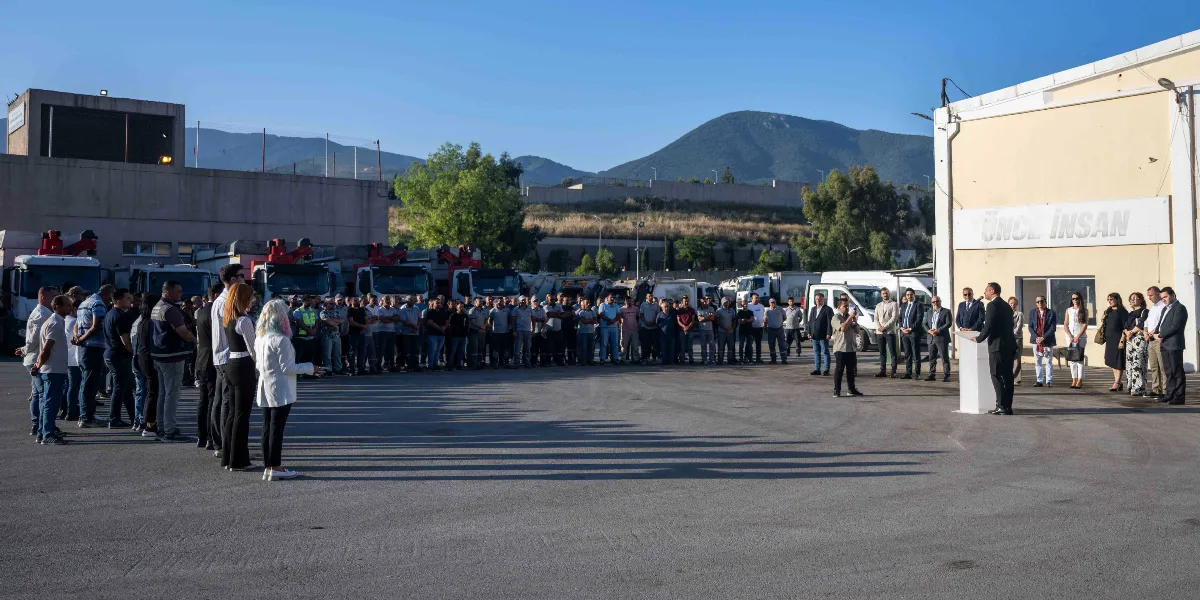 Açık havada, Bornova Belediyesi şantiye alanında toplanmış kalabalık bir grup çalışan ve yetkili, bayramlaşma töreni için bir araya gelmiş. Arka planda belediyeye ait çöp kamyonları ve dağ manzarası yer alırken, sağ taraftaki binanın üzerinde 