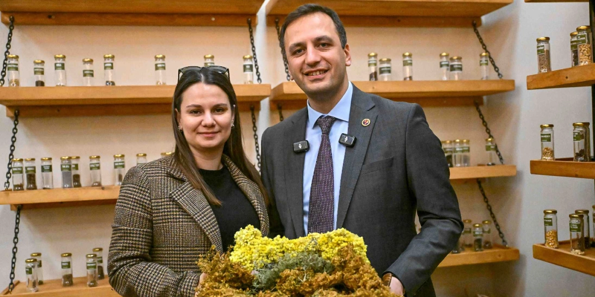 Koyu renkli ekose bir ceket giyen kadın ile lacivert takım elbiseli ve kravatlı bir erkek, arkalarında yerel tohumların bulunduğu cam kavanozlarla dolu ahşap rafların önünde yan yana duruyor. İkili, ellerinde tuttukları büyük bir demet sarı ve yeşil bitkiyle objektife gülümseyerek poz veriyor.